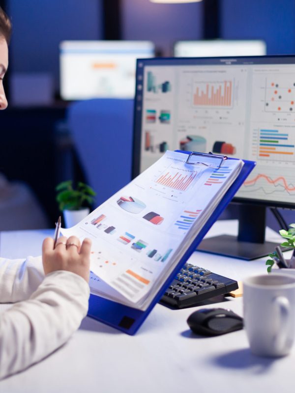Freelancer woman comparing graphics from clipboard with grafics from computer at business office. Tired focused employee using modern technology network wireless working on project solution